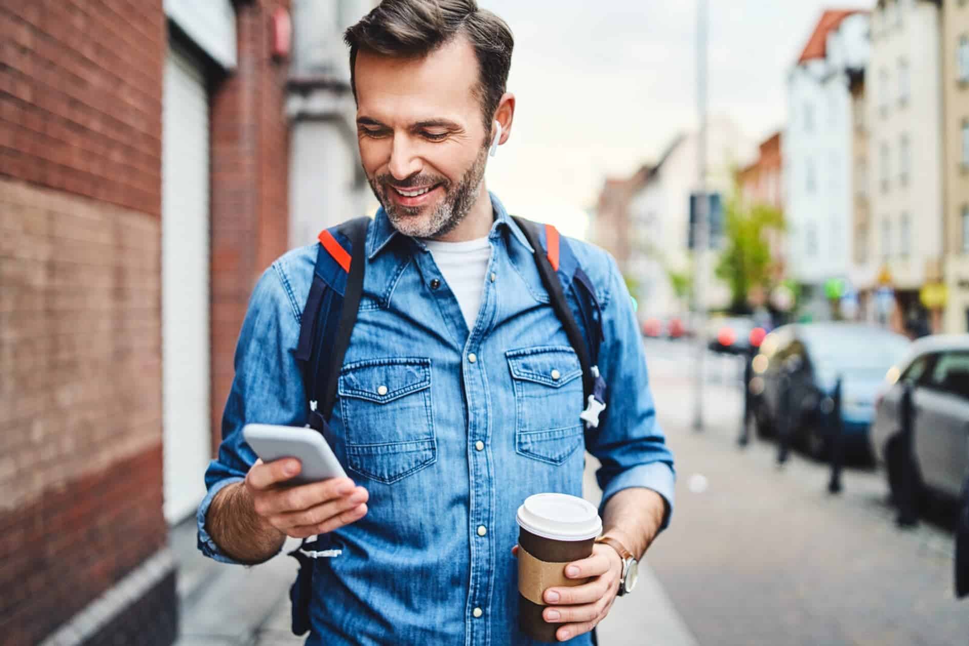 A man walking down the pavement with a takeaway coffee in one hand and his mobile phone in the other.