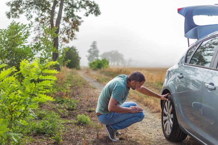 Crouched man looking at the wheel of his car on rural road with fog