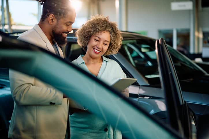 Happy woman using digital tablet with a salesman while buying new car in showroom