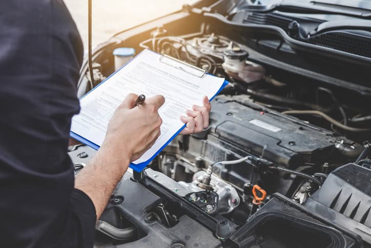 a close up of a mechanic with a clipboard looking into the bonnet of a car
