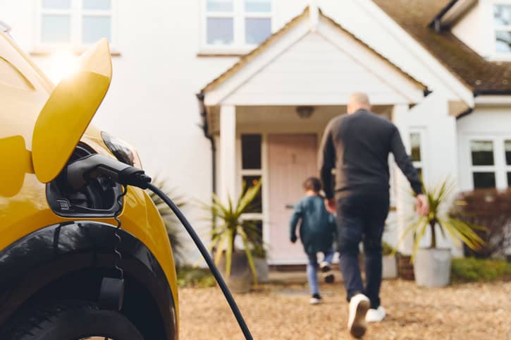 A close up of a yellow electric car charging on a family drive with a parent and child in the background